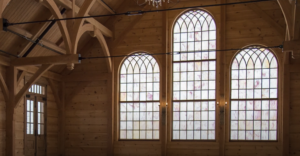 A spacious wooden interior with arched beams, three large tall windows with grid patterns, and a chandelier hanging from the ceiling—an ideal setting for creative marketing or advertising shoots, where natural light highlights the stunning woodwork.