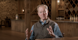 An older man sits indoors at a wooden table, gesturing as he discusses advertising strategies. Shelves with wine bottles, wooden beams, and warm lighting behind him create a rustic, cozy atmosphere.