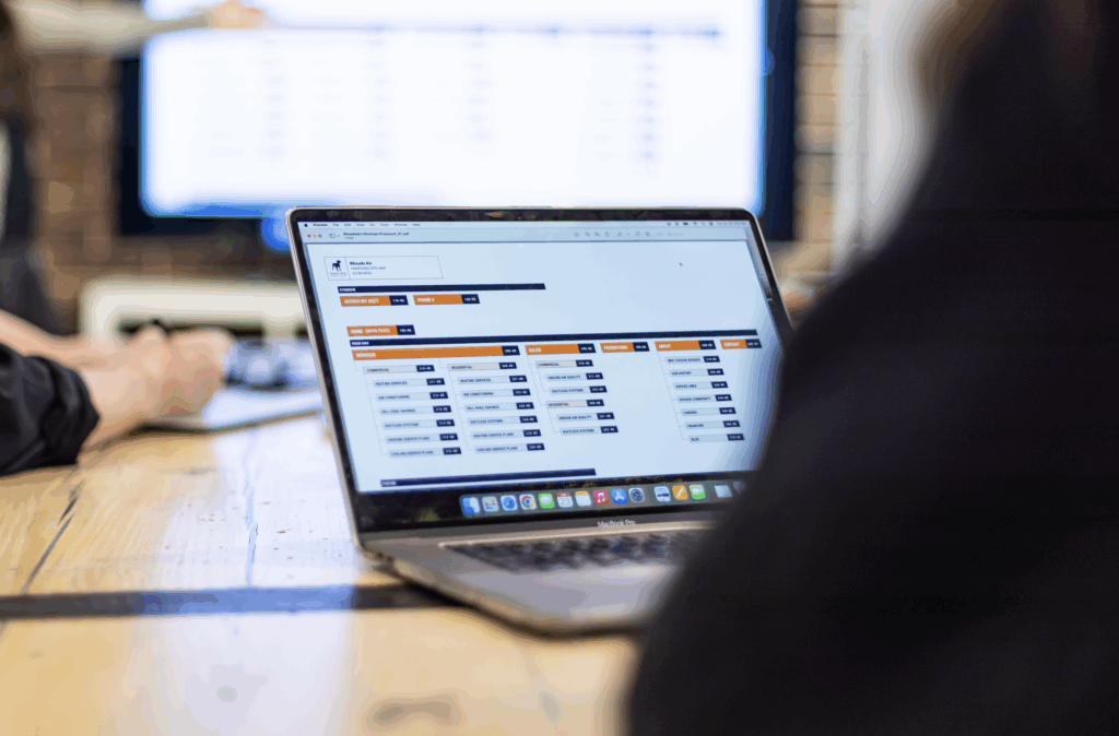 A laptop on a wooden table displays a spreadsheet or data management application, perfect for marketing analytics. In the background, two people work together, with a large screen showing a blurred table or chart.