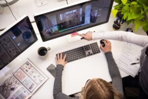 Two people work at a desk with a computer, reviewing interior design images on the monitor. One person points at the screen while the other uses a mouse. Papers, a magazine, and a coffee cup are nearby.