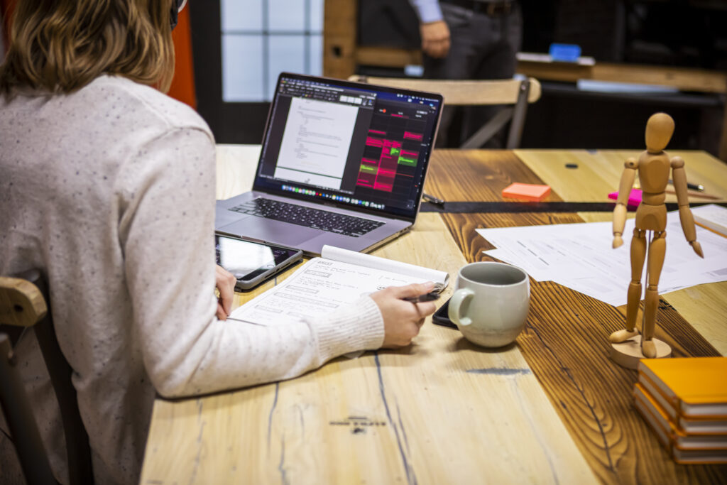 A person sits at a wooden table working on a laptop with documents, a mug, a phone, and a wooden mannequin nearby. The laptop displays charts and text, and notebooks are stacked on the table.