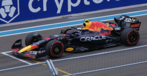 A Red Bull Racing Formula 1 car, featuring Oracle and other sponsor logos, is parked on a racetrack near a blue wall with the Crypto.com logo. The driver is wearing a helmet inside the car.