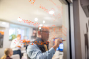 A person writes on a glass wall with a red marker in an office setting, with handwritten notes and diagrams visible on the glass. The background shows blurred people and office equipment.