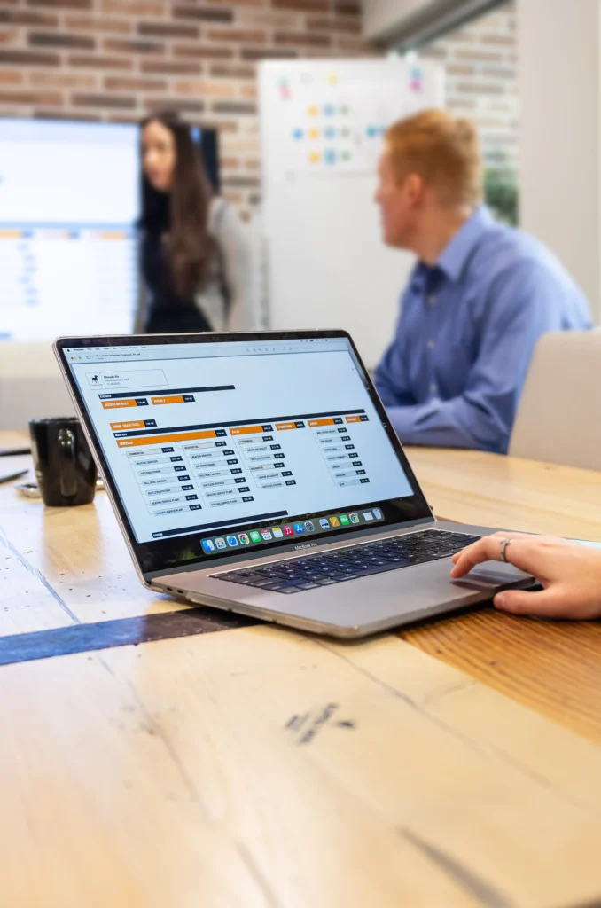 A person uses a laptop displaying a spreadsheet at a wooden table. In the background, two people sit and talk near a large screen in a modern office setting.