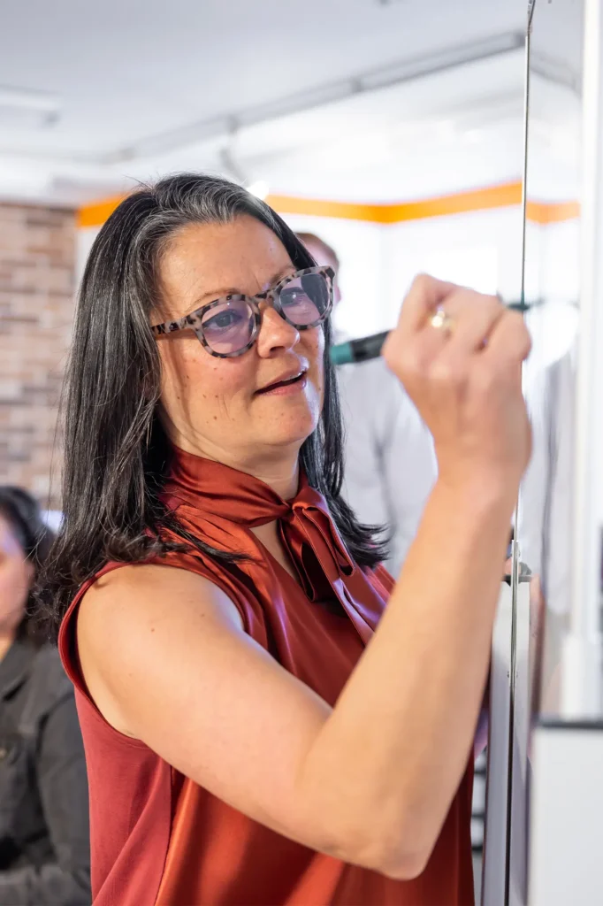A woman with glasses and long dark hair writes on a whiteboard with a marker. She is wearing a sleeveless red blouse and is focused on her work, with other people blurred in the background.