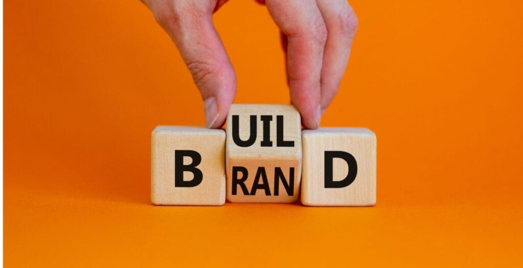 A hand arranges wooden blocks on an orange background to spell BUILD BRAND, with one block showing both RAN and the letter I partially displayed.