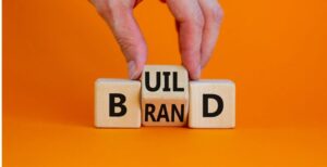 A hand arranges wooden blocks on an orange background to spell BUILD BRAND, with one block showing both RAN and the letter I partially displayed.