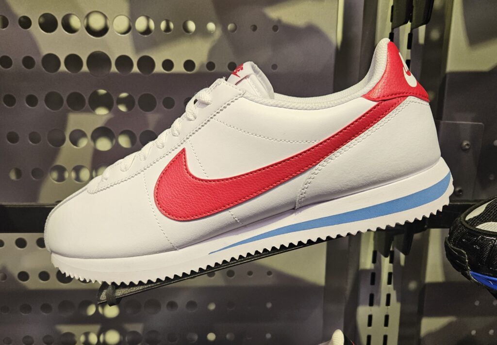 A white Nike sneaker with a red swoosh logo, red heel tab, and blue stripe on the white sole, displayed on a perforated metal wall in a store.