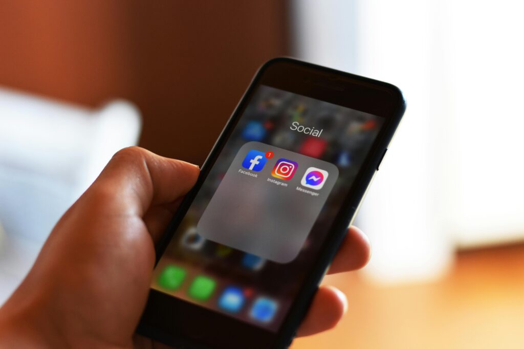 A hand holding a smartphone displaying a folder labeled Social with Facebook, Instagram, and Messenger app icons on the screen. The background is blurred.