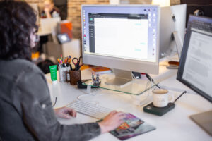 Person with curly hair using a computer at a desk, with various office supplies, a mug, and another monitor nearby. Background shows another person working and shelves with items.