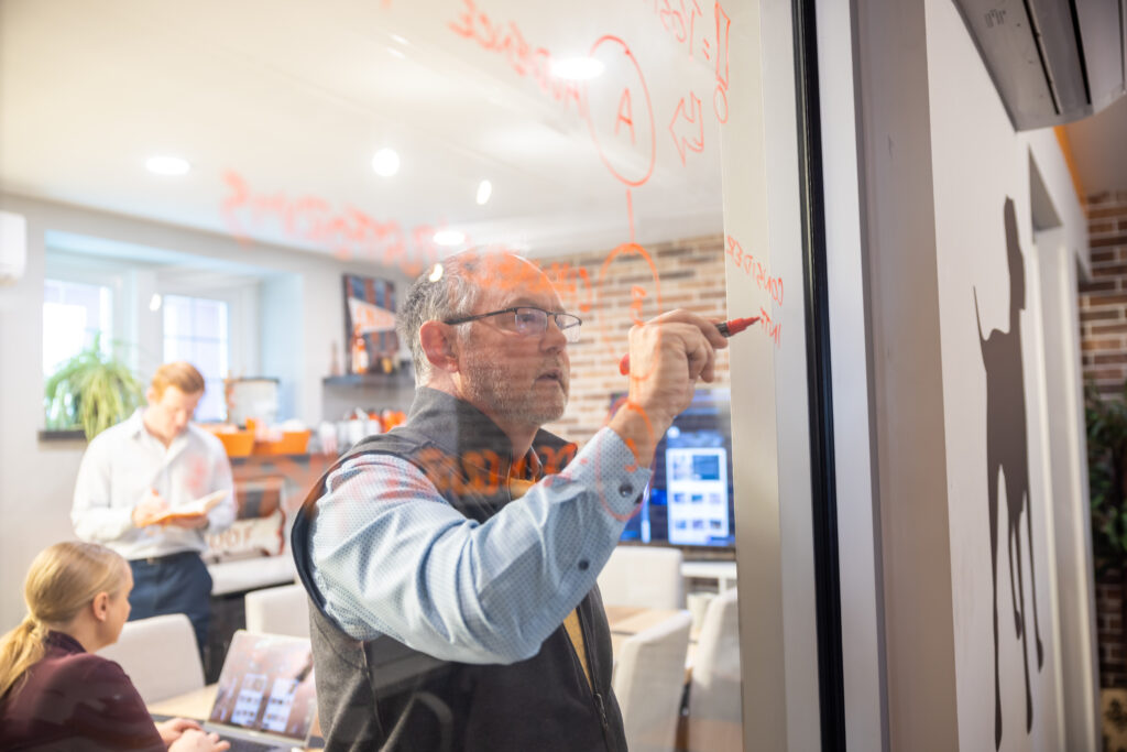 Man writes on a glass wall with a red marker in a modern office, while colleagues work at a table behind him.