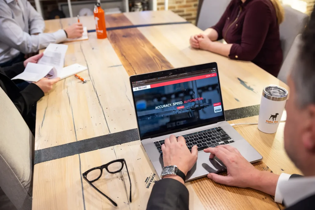 A person uses a laptop displaying the Flex/cell website at a wooden meeting table with colleagues.