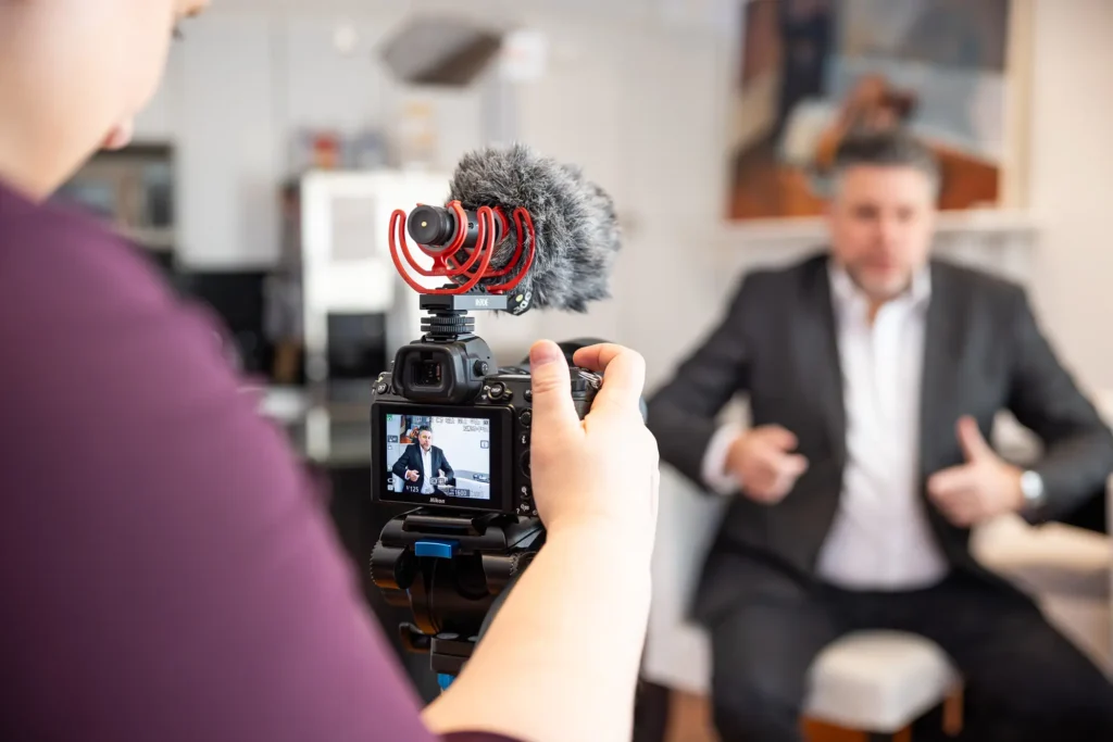 A person operates a camera on a tripod with a microphone to record a man in a suit sitting across from them.